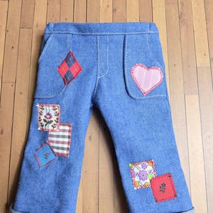 Peut inclure: Pantalon en jean bleu orné de divers empiècements en tissu coloré cousus sur le devant. Les pièces incluent un losange rouge, un cœur rose et plusieurs rectangles à motifs floraux, à carreaux et rouge uni avec des coutures apparentes.