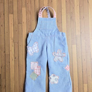 May include: Light blue denim overalls featuring a bib pocket, two side pockets, and adjustable straps with a pink and white gingham pattern. The garment is decorated with various fabric patches including a butterfly, flowers, and squares.