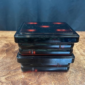 May include: Two stacked, square black lacquer boxes with red detailing. The top box features six red dots. The boxes are on a wooden surface. The boxes have a glossy finish and a traditional design.