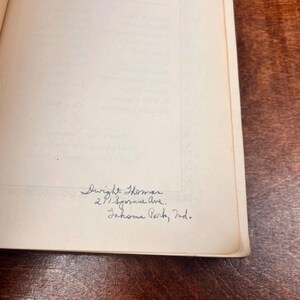 以下が含まれることがあります： 青いインクで手書きされたテキストが書かれた開いた本。テキストは「Dwight Thomas, 291 Spruce Ave., Inkoma Park, Ind.」と書かれています。本のページはオフホワイトで、背景は濃い木材の表面です。
