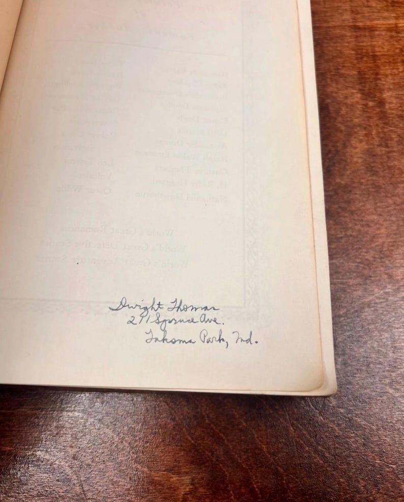 以下が含まれることがあります： 青いインクで手書きされたテキストが書かれた開いた本。テキストは「Dwight Thomas, 291 Spruce Ave., Inkoma Park, Ind.」と書かれています。本のページはオフホワイトで、背景は濃い木材の表面です。