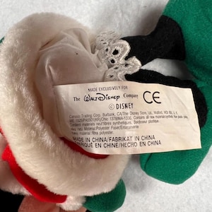 K&ouml;nnte beinhalten: Ein Stofftier mit einem wei&szlig;en Hut mit rotem Rand, schwarzen und gr&uuml;nen Akzenten und einem Etikett mit der Aufschrift "MADE EXCLUSIVELY FOR The Walt Disney Company". Das Spielzeug hat ein CE-Zeichen und das Disney-Logo.
