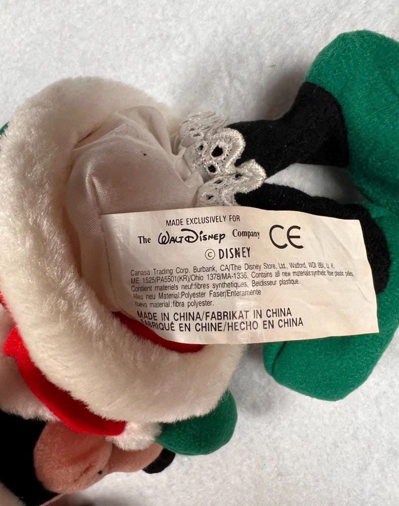 K&ouml;nnte beinhalten: Ein Stofftier mit einem wei&szlig;en Hut mit rotem Rand, schwarzen und gr&uuml;nen Akzenten und einem Etikett mit der Aufschrift "MADE EXCLUSIVELY FOR The Walt Disney Company". Das Spielzeug hat ein CE-Zeichen und das Disney-Logo.