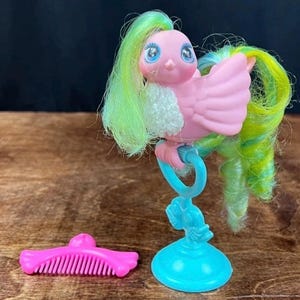 Könnte beinhalten: Ein Vintage-Spielzeugvogel mit rosa Flügeln, blauen Augen und grün-gelbem Haar. Der Vogel sitzt auf einem blauen Ständer mit einem passenden rosa Kamm. Das Spielzeug hat eine Retro-Ästhetik.