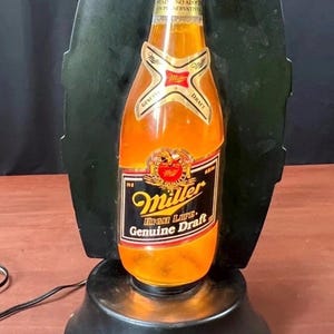 May include: A Miller Genuine Draft beer bottle shaped electric lamp with a gold cap, orange glowing body, and black base. The label features the Miller logo, the text High Life, and Genuine Draft. It stands against a black vertical backing.