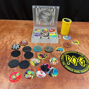 Puede incluir: Una colección de POG, que incluye una caja de plástico transparente con POG variados, un tubo amarillo y un tablero de juego negro con texto amarillo. Los POG presentan varios diseños coloridos, incluyendo calaveras, personajes de dibujos animados y patrones abstractos.