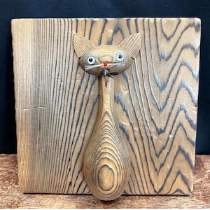 May include: Wooden cat sculpture mounted on a wooden plank. The cat has a round head with large ears, round eyes, and a red nose. The body is elongated, resembling a spoon. The wood grain is visible throughout the piece.