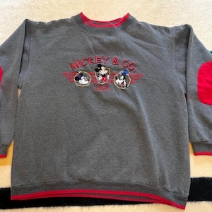 May include: Gray sweatshirt with red accents and embroidered "Mickey & Co." logo featuring Mickey Mouse, Donald Duck, and Goofy. The sweatshirt has red elbow patches and trim around the collar, cuffs, and hem.
