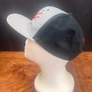 Puede incluir: Gorra de b&eacute;isbol negra y gris con visera plana. La gorra presenta un panel blanco con motas negras y letras rojas bordadas. La parte trasera de la gorra es negra y la visera es gris.