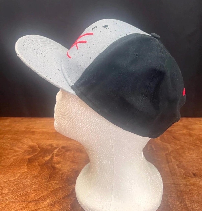 Puede incluir: Gorra de b&eacute;isbol negra y gris con visera plana. La gorra presenta un panel blanco con motas negras y letras rojas bordadas. La parte trasera de la gorra es negra y la visera es gris.