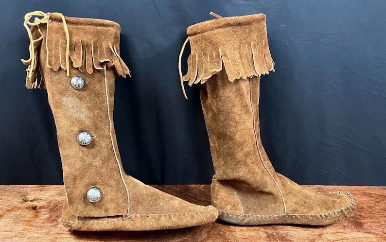 May include: A pair of knee-high brown suede boots with a fringed top and decorative silver buttons. The boots have a moccasin-style sole and are displayed on a wooden surface against a dark background.
