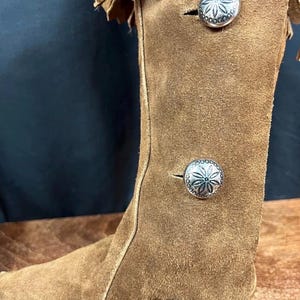 May include: A brown suede boot with decorative silver buttons and fringe detailing. The boot has a tall shaft and a classic Western-inspired design, perfect for adding a touch of rustic charm to any outfit.