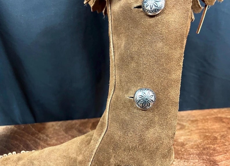 May include: A brown suede boot with decorative silver buttons and fringe detailing. The boot has a tall shaft and a classic Western-inspired design, perfect for adding a touch of rustic charm to any outfit.