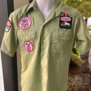 May include: Olive green short-sleeved work shirt with multiple Texaco patches. Patches include the Texaco logo, a truck, and the words "National Truck Roadeo 1979" and "National Safety Services". The shirt has two chest pockets and a button-down front.