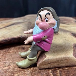 May include: A Grumpy dwarf figurine featuring a pink tunic with a patch on the sleeve, a brown hat, dark gray trousers, and light green pointed shoes. The character has a large pink nose, a white beard, and a frowning expression with one eye closed.