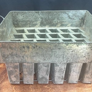 Może przedstawiać: Zabytkowa, prostokątna metalowa forma do lodu z uchwytem. Forma ma siatkę prostokątnych przegródek i jest podparta pionowymi prętami. Metal ma zwietrzały wygląd, co sugeruje, że jest to antyczny przedmiot kuchenny.