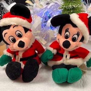 K&ouml;nnte beinhalten: Zwei Pl&uuml;schpuppen von Mickey Mouse und Minnie Mouse in Weihnachtsmann-Outfits. Jede Puppe tr&auml;gt eine rote Jacke mit wei&szlig;em Besatz, einen schwarzen G&uuml;rtel und einen Weihnachtsmannhut. Sie haben gr&uuml;ne Handschuhe und Schuhe.