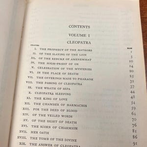 以下が含まれることがあります： 「クレオパトラ」第1巻の目次ページが表示された開いた本。ページには、「ハトホル神の予言」や「ライオンの殺害」などの章タイトルが記載されています。本のページは古く、テキストが見えます。