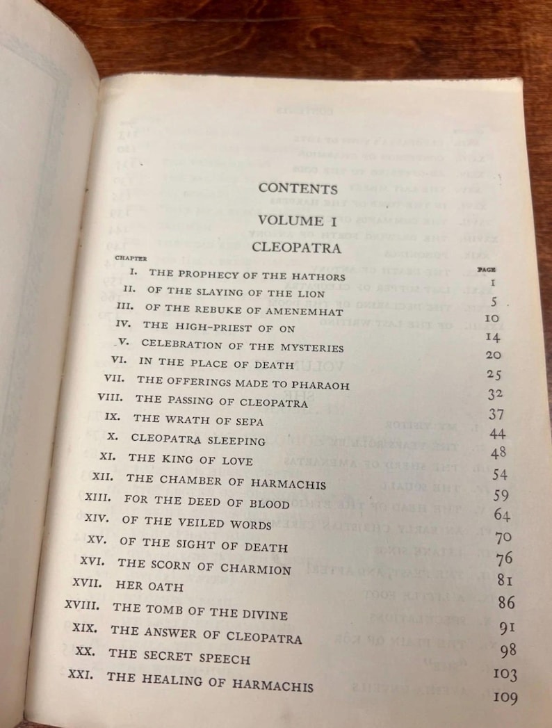 以下が含まれることがあります： 「クレオパトラ」第1巻の目次ページが表示された開いた本。ページには、「ハトホル神の予言」や「ライオンの殺害」などの章タイトルが記載されています。本のページは古く、テキストが見えます。