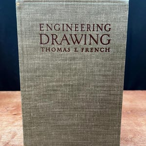 May include: A vintage book titled "Engineering Drawing" by Thomas E. French. The book has a textured, olive-green cover with the title and author's name in maroon lettering. The book is standing upright on a wooden surface.