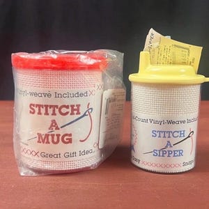 K&ouml;nnte beinhalten: Ein Stitch A Mug Set mit rotem Deckel in transparenter Kunststoffverpackung und ein Stitch A Sipper Set mit gelbem Deckel. Beide Artikel haben wei&szlig;e Gitteroberfl&auml;chen f&uuml;r Handarbeiten, blauen Text und rote Kreuzstichmuster auf den Etiketten.