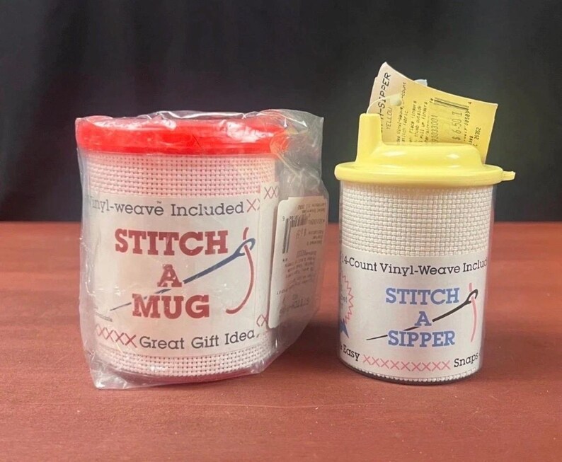 K&ouml;nnte beinhalten: Ein Stitch A Mug Set mit rotem Deckel in transparenter Kunststoffverpackung und ein Stitch A Sipper Set mit gelbem Deckel. Beide Artikel haben wei&szlig;e Gitteroberfl&auml;chen f&uuml;r Handarbeiten, blauen Text und rote Kreuzstichmuster auf den Etiketten.