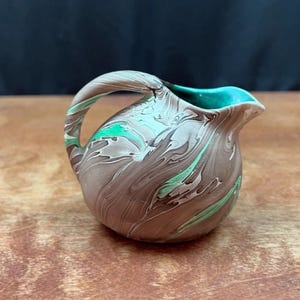May include: A small, decorative jug with a handle. The jug features a brown, grey, and white marbled design with teal accents on the rim and green accents throughout. The jug is sitting on a wooden surface.