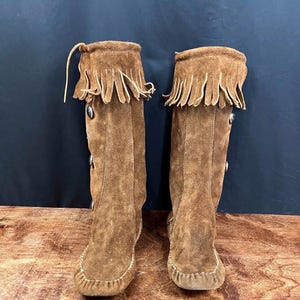 May include: A pair of knee-high brown suede boots with fringe detailing at the top. The boots have decorative buttons and a moccasin-style toe. The boots are displayed on a wooden surface against a dark background.