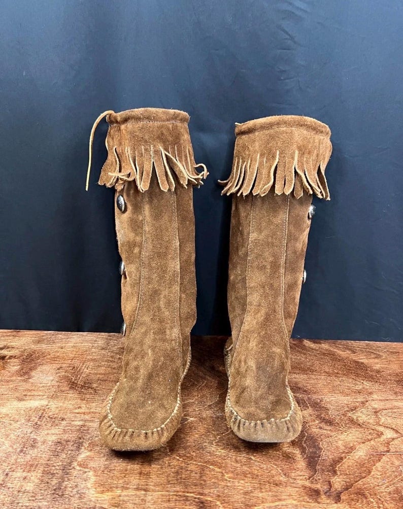May include: A pair of knee-high brown suede boots with fringe detailing at the top. The boots have decorative buttons and a moccasin-style toe. The boots are displayed on a wooden surface against a dark background.