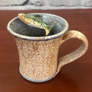 May include: A ceramic mug with a speckled tan and brown exterior and a light blue interior. A small, three-dimensional green and yellow fish figure is attached to the inner rim, appearing to swim across the top of the mug's opening.