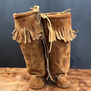 May include: A pair of brown suede boots with fringed tops and leather ties. The boots have a moccasin-style construction with stitched soles. The boots are approximately 12 inches tall.