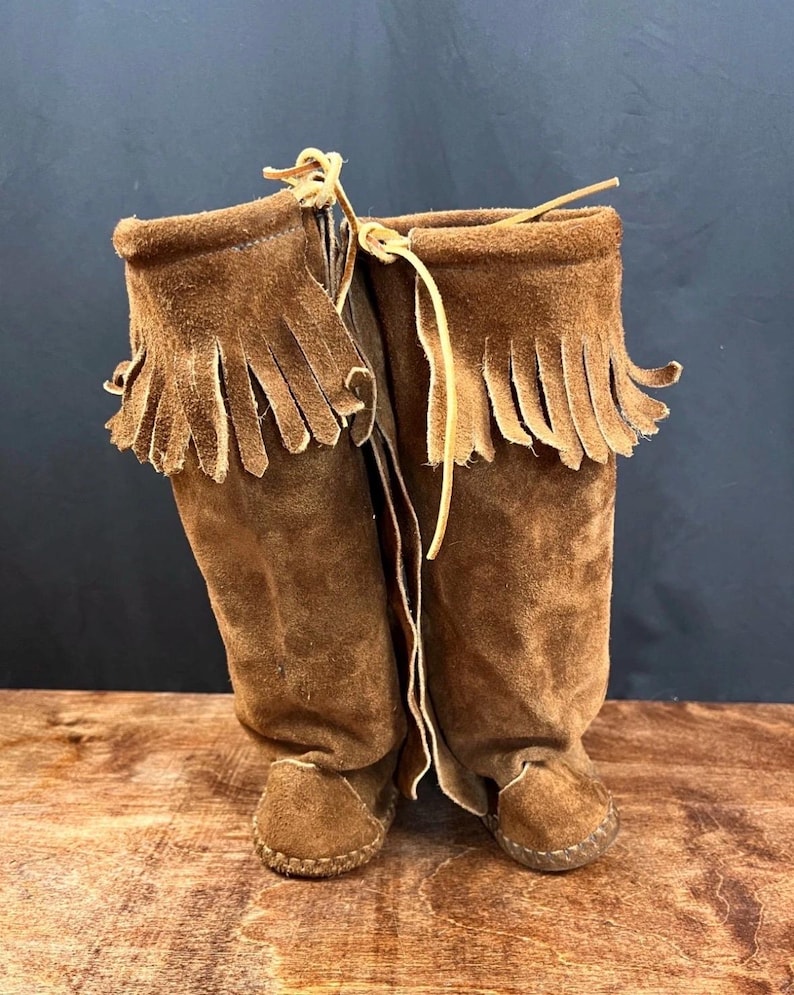 May include: A pair of brown suede boots with fringed tops and leather ties. The boots have a moccasin-style construction with stitched soles. The boots are approximately 12 inches tall.
