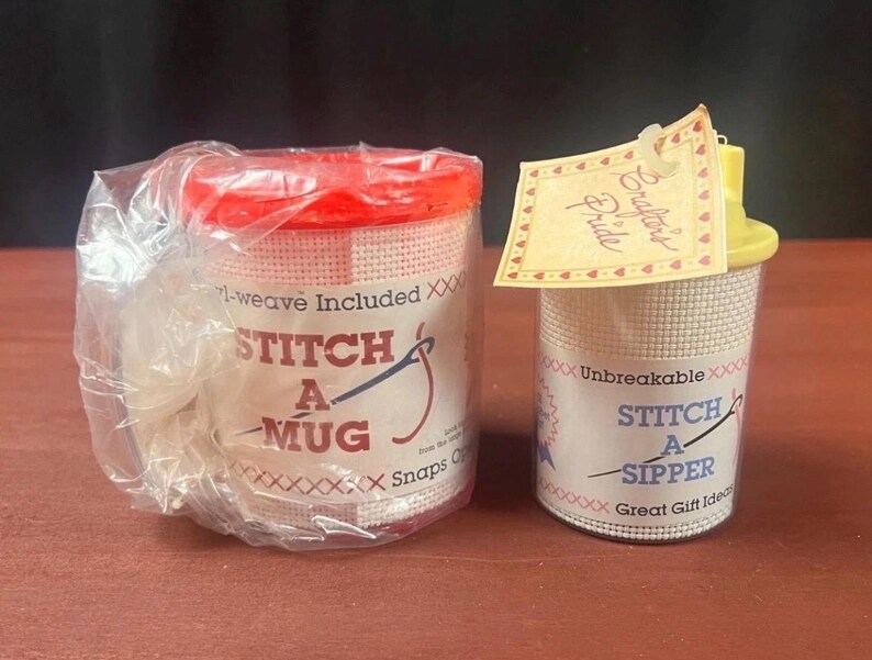 K&ouml;nnte beinhalten: Zwei Kunststoff-Bastelbeh&auml;lter mit wei&szlig;er Netzau&szlig;enseite f&uuml;r Handarbeiten. Der linke Beh&auml;lter hat einen roten Deckel und ein Etikett mit der Aufschrift STITCH A MUG. Der rechte hat einen gelben Deckel, ein Crafter's Pride-Etikett und die Aufschrift STITCH A SIPPER.