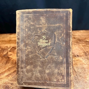 以下が含まれることがあります： 茶色の革装丁のヴィンテージ本で、表紙に「H. Rider Haggard」のタイトルがエンボス加工されています。 本には、著者の名前が装飾的なフォントで書かれた盾のデザインがあります。 表紙には使用感があり、クラシックでアンティークな外観になっています。