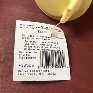 K&ouml;nnte beinhalten: Ein gelber Kunststoffdeckel mit Ausguss, befestigt an einem wei&szlig;en rechteckigen Produktetikett. Das Etikett tr&auml;gt die Aufschrift STITCH-A-SIPPER YELLOW, enth&auml;lt Vinyl-Webstoff f&uuml;r Kreuzstich, &Ouml;ffnungsanweisungen, Sp&uuml;lmaschinenfestigkeit und Barcode.