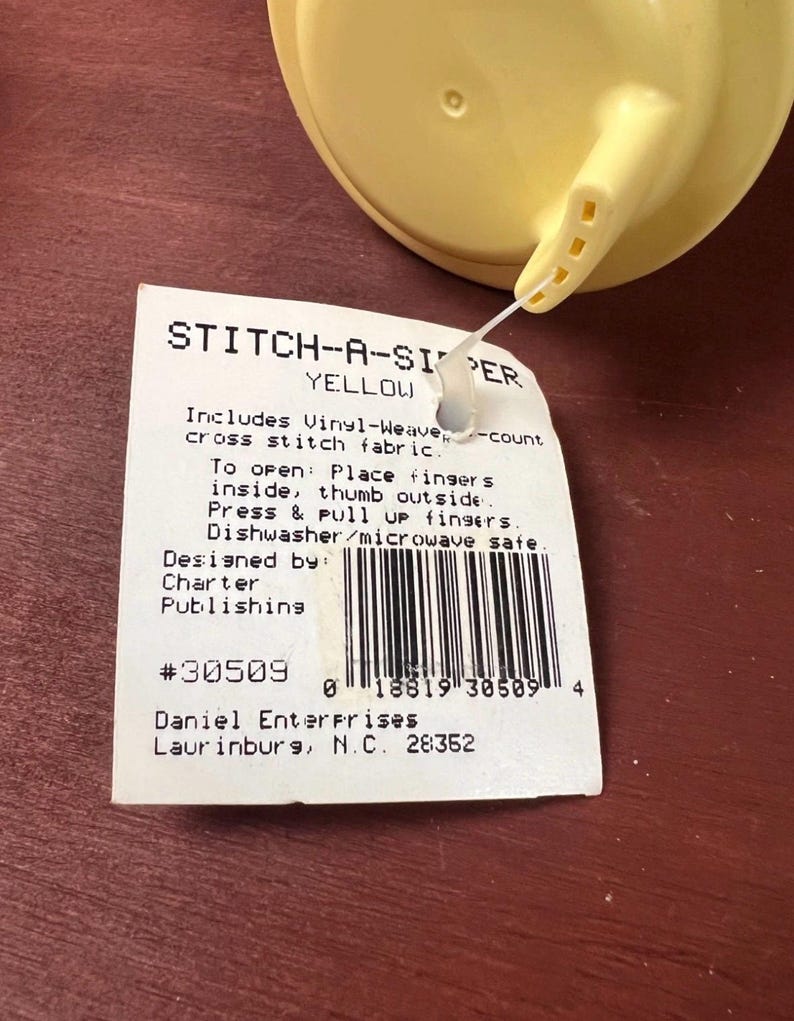 K&ouml;nnte beinhalten: Ein gelber Kunststoffdeckel mit Ausguss, befestigt an einem wei&szlig;en rechteckigen Produktetikett. Das Etikett tr&auml;gt die Aufschrift STITCH-A-SIPPER YELLOW, enth&auml;lt Vinyl-Webstoff f&uuml;r Kreuzstich, &Ouml;ffnungsanweisungen, Sp&uuml;lmaschinenfestigkeit und Barcode.
