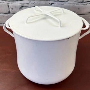 May include: A solid white pot with a matching lid and two side handles. The lid features a distinct cross-shaped handle on top. The pot has a smooth, rounded body and a wide rim, presenting a clean, uniform appearance throughout the entire structure.
