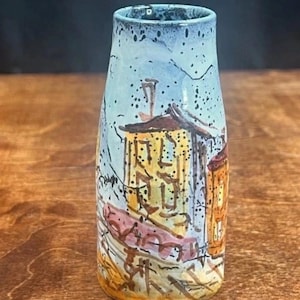 May include: A hand-painted ceramic vase with a cityscape design. The vase features a light blue top and a body adorned with yellow, orange, and brown buildings. Black lines and splatters add detail to the artwork. The vase is on a wooden surface.