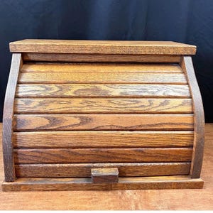 May include: A medium brown wooden bread box featuring a curved roll-top door with horizontal slats. The container has a flat rectangular top surface, a solid base, and a small rectangular handle attached to the bottom edge of the sliding door.