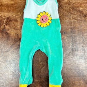 Pode incluir: Macacão de uma peça para bebé em tecido macio e felpudo. A parte superior é branca, o meio é verde brilhante e os pés são amarelos. Uma aplicação de girassol sorridente está centrada no peito. O macacão tem fechos de pressão nos ombros.