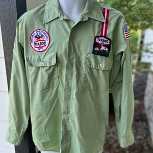 May include: Light green long-sleeve shirt with two chest pockets. Features Texaco and National Truck Roadeo 1971 patches. A red and white striped accent is on the right shoulder. The shirt is displayed on a mannequin.
