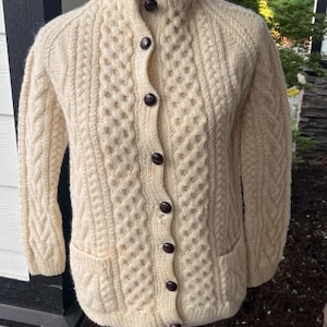 May include: A cream-colored cable knit cardigan featuring a high collar, long sleeves, and a front button closure with dark brown buttons. The garment includes two front pockets and a textured pattern of vertical cables and honeycomb stitches.