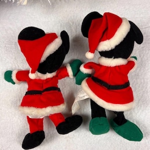 K&ouml;nnte beinhalten: Zwei Pl&uuml;schfiguren in Weihnachtsmann-Outfits. Die Figuren haben schwarze K&ouml;rper, rote Jacken und H&uuml;te mit wei&szlig;em Besatz, gr&uuml;ne Handschuhe und Schuhe. Die Figuren halten sich an den H&auml;nden und sind bereit f&uuml;r die Feiertage.