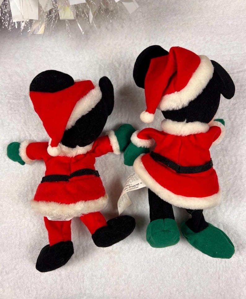 K&ouml;nnte beinhalten: Zwei Pl&uuml;schfiguren in Weihnachtsmann-Outfits. Die Figuren haben schwarze K&ouml;rper, rote Jacken und H&uuml;te mit wei&szlig;em Besatz, gr&uuml;ne Handschuhe und Schuhe. Die Figuren halten sich an den H&auml;nden und sind bereit f&uuml;r die Feiertage.