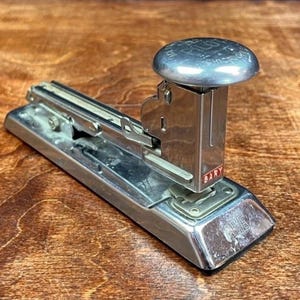 May include: A vintage silver stapler with a rounded top and a rectangular base. The stapler has the word "GARY" printed on the side in red. The stapler is on a wooden surface.