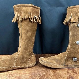 May include: A pair of knee-high brown suede boots. Each boot features a fringed top, decorative silver buttons, and a moccasin-style sole. The boots are displayed on a wooden surface against a dark blue background.