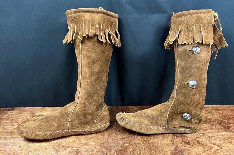 May include: A pair of knee-high brown suede boots. Each boot features a fringed top, decorative silver buttons, and a moccasin-style sole. The boots are displayed on a wooden surface against a dark blue background.