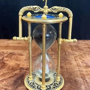 Puede incluir: Un reloj de arena ornamentado con un marco de metal dorado. El reloj de arena tiene una bombilla de vidrio transparente en la parte inferior y una bombilla llena de arena negra en la parte superior. La parte superior e inferior tienen patrones decorativos y detalles azules. Altura: aprox. 15 cm.