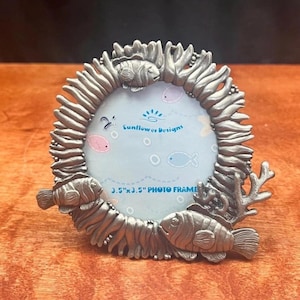 May include: A silver-colored photo frame with a sea life theme. The frame is round with a textured border and features three fish and coral accents. The frame holds a 3.5" x 3.5" photo and has the text "Sunflower Designs".
