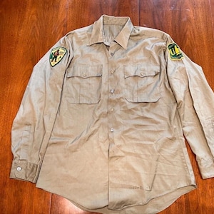 Op de afbeelding: Beige overhemd met lange mouwen, twee borstzakken en patches op beide mouwen. De patches hebben groene en gele accenten. Het overhemd is gemaakt van een lichtgewicht materiaal.